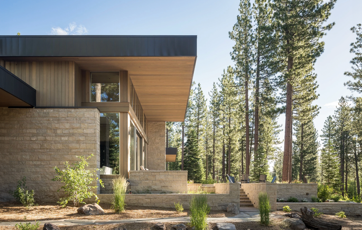 Modern single-story house with a dark roof overhang, light wood siding, and stone masonry, set among tall pine trees with landscaped grounds.