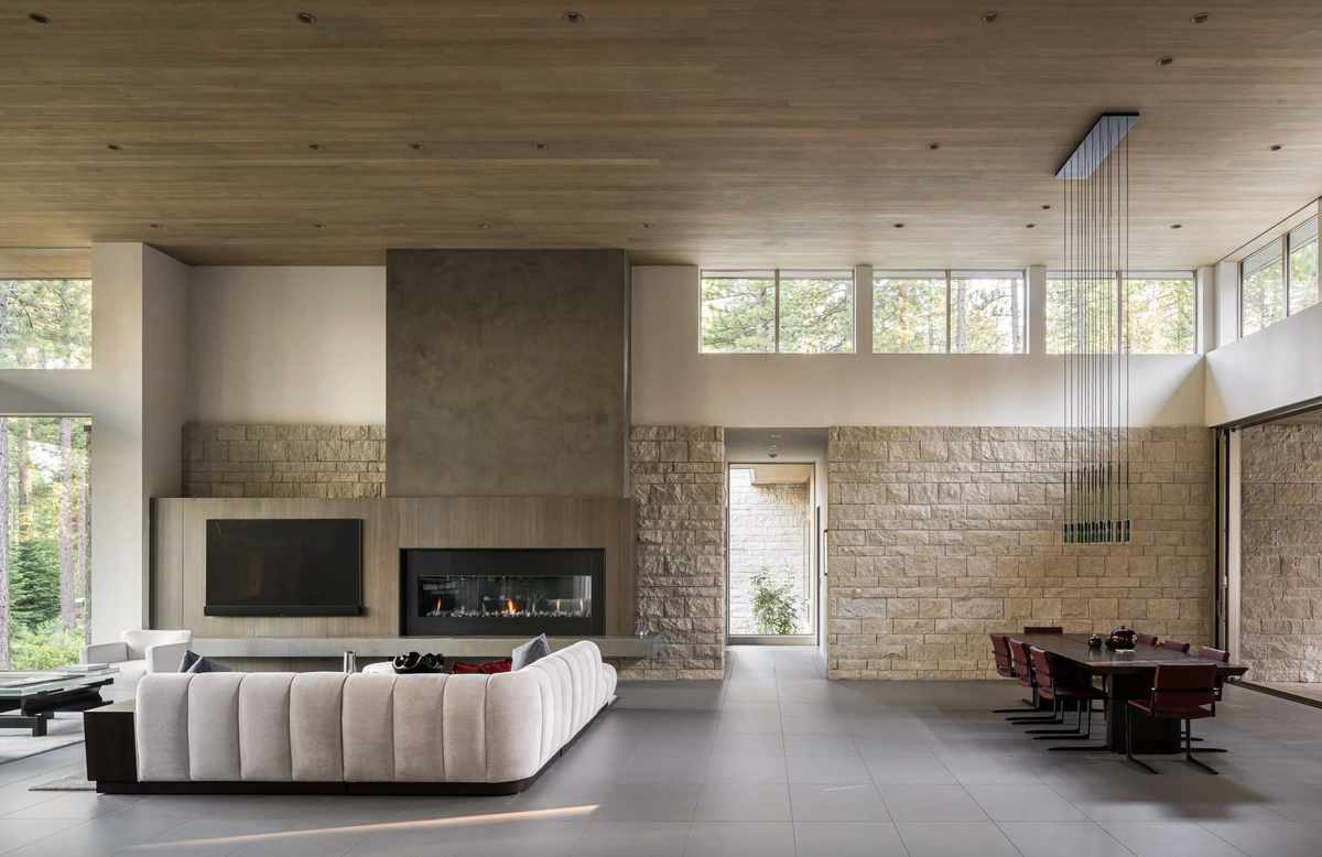A modern open-plan living space with a low, tufted cream sectional sofa in the foreground, a stone and wood fireplace, high wood ceiling, dining area with a dark wood table and chairs, and tall windows looking out to trees.