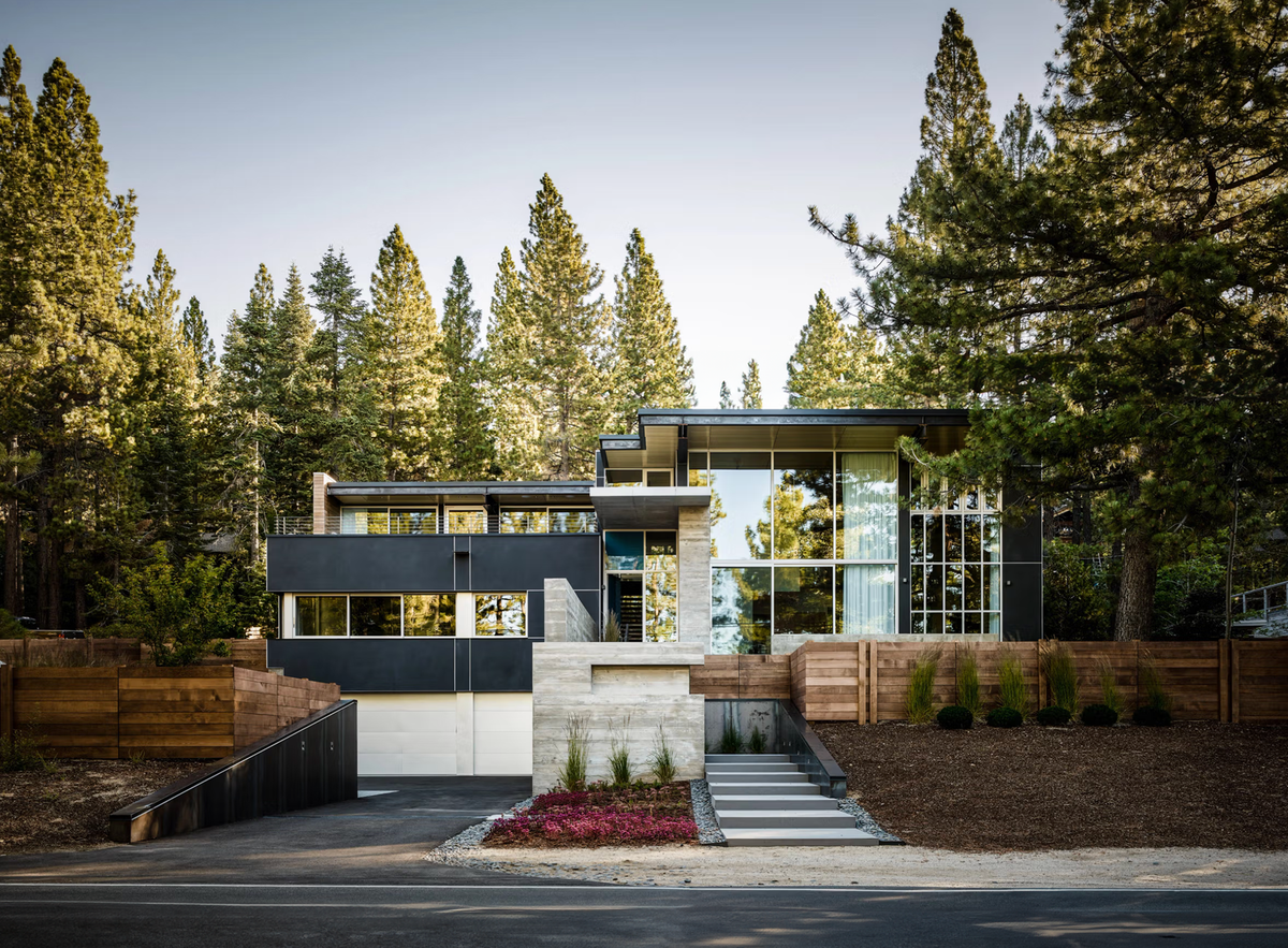 Modern single-story house with large glass walls and dark exterior panels, set among tall evergreen trees with a driveway and landscaped entrance in the foreground.