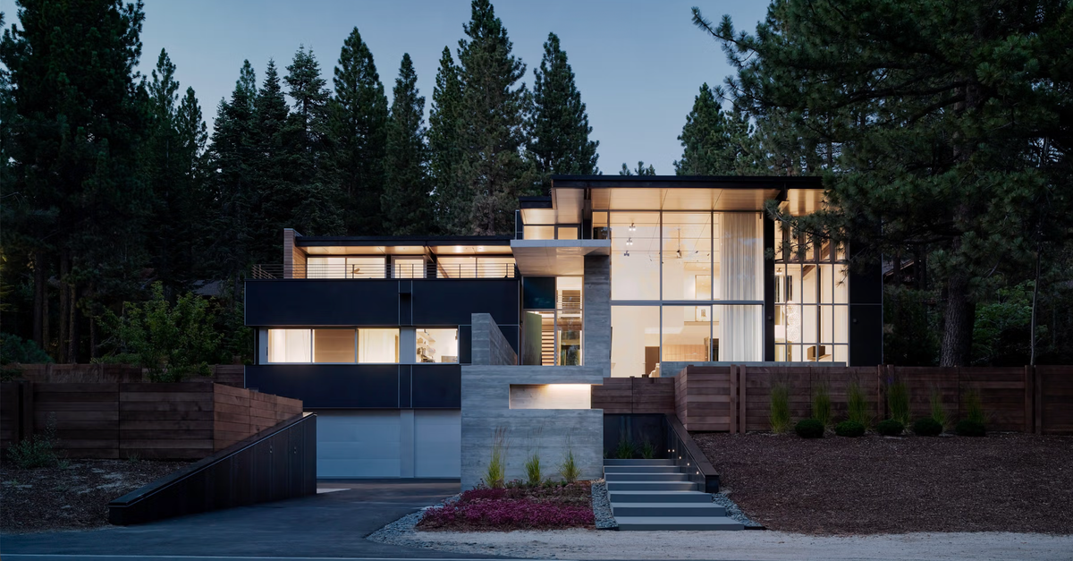 Modern single-story house with large glass walls and warm interior lighting, set among tall evergreen trees at dusk. The facade includes dark charcoal and light gray sections, concrete stairs, and landscaped wood retaining walls.