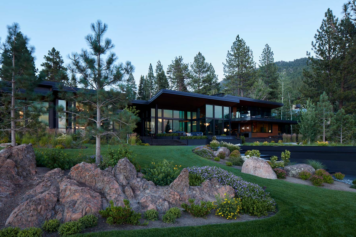 Modern single-story house with large glass walls and dark overhanging roofs, set in a landscaped lawn with rocks, shrubs, and tall pine trees at dusk.