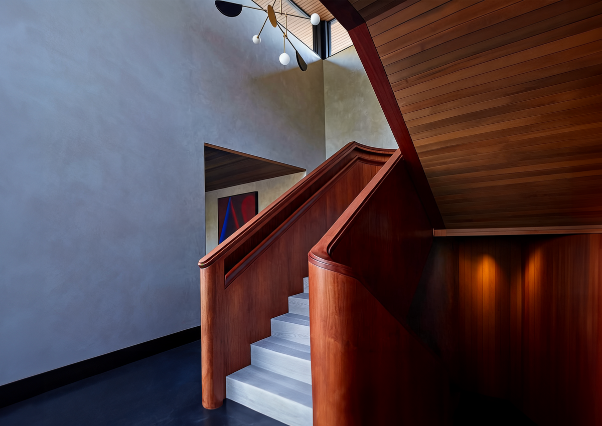 A modern interior staircase with a rich reddish-brown wooden handrail and matching wood ceiling, featuring white steps and a textured gray plaster wall.